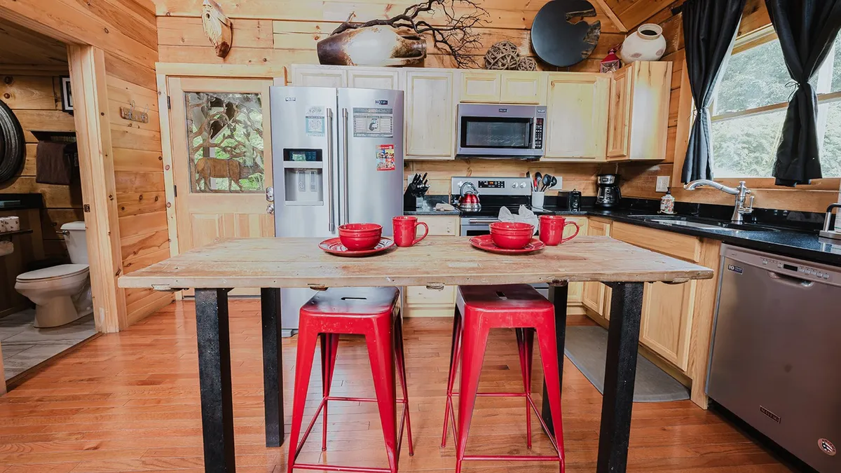 This image features a wooden dining table with red stools, modern kitchen appliances, and charming decorative touches on the walls. The setup is perfect for both cooking and socializing, reflecting a cozy dining environment where guests can enjoy meals and each other's company.