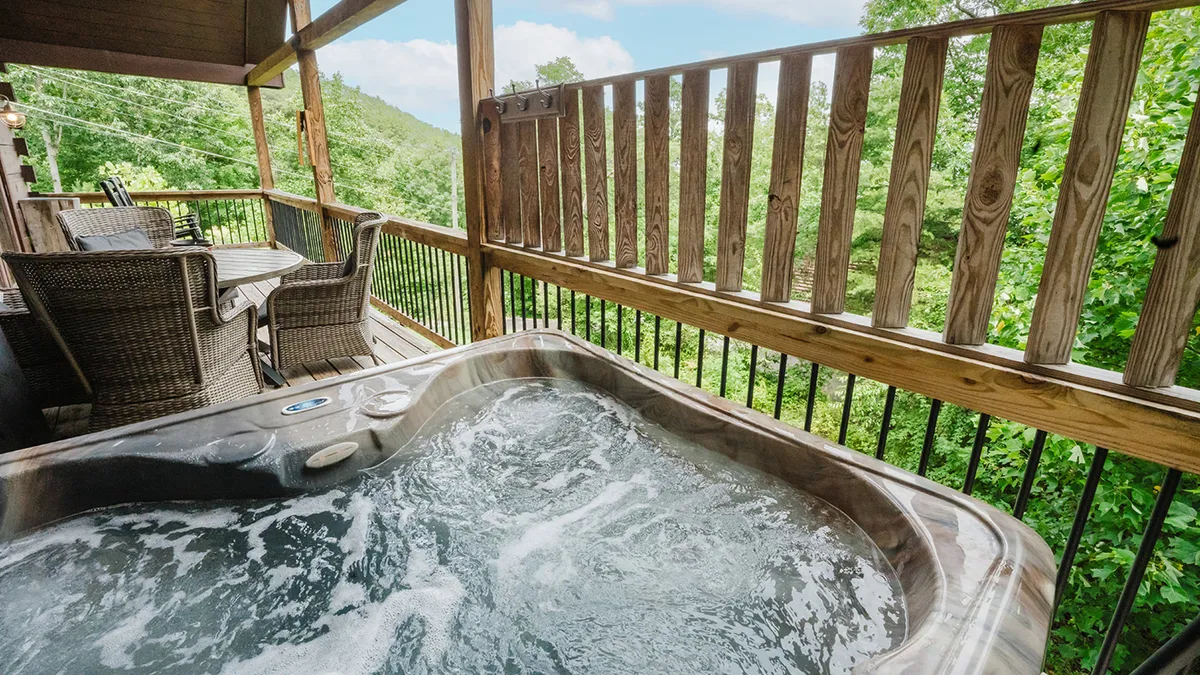 This image captures an inviting covered deck featuring a private hot tub, wooden railing, and an outdoor dining setup. The serene setting encourages relaxation and socializing, with the hot tub positioned to take full advantage of the surrounding natural beauty. Sunlight filters through the trees, casting a soft glow over the space, and the setup hints at the possibility of both early morning coffee and evening barbecues.