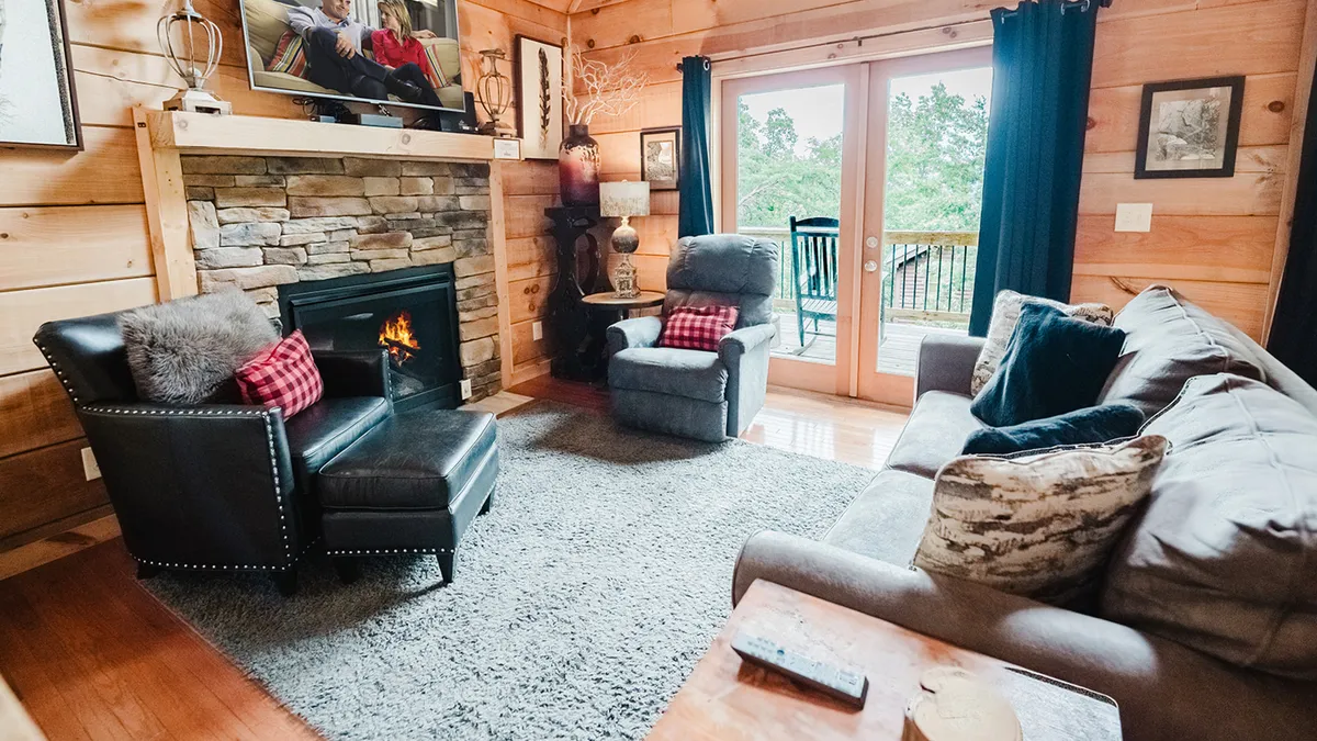 This image presents a cozy interior with a fireplace, large windows, and comfortable seating arranged for socializing. The warmth of the fire is complemented by natural light streaming in, evoking the inviting atmosphere of a mountain retreat designed for connection.