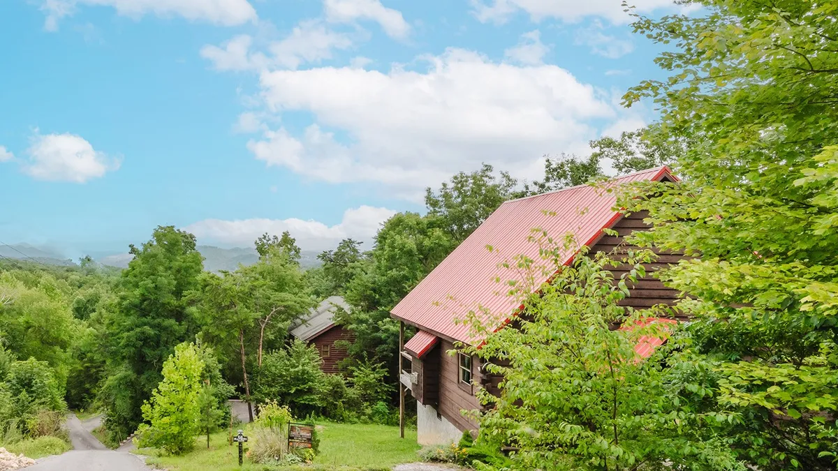 This image shows a charming wooden cabin with a red roof nestled among lush green trees, with a dirt path leading up to the entrance. The setting suggests privacy and immersion in nature, perfect for families looking to explore the Smoky Mountains and return to a peaceful retreat.