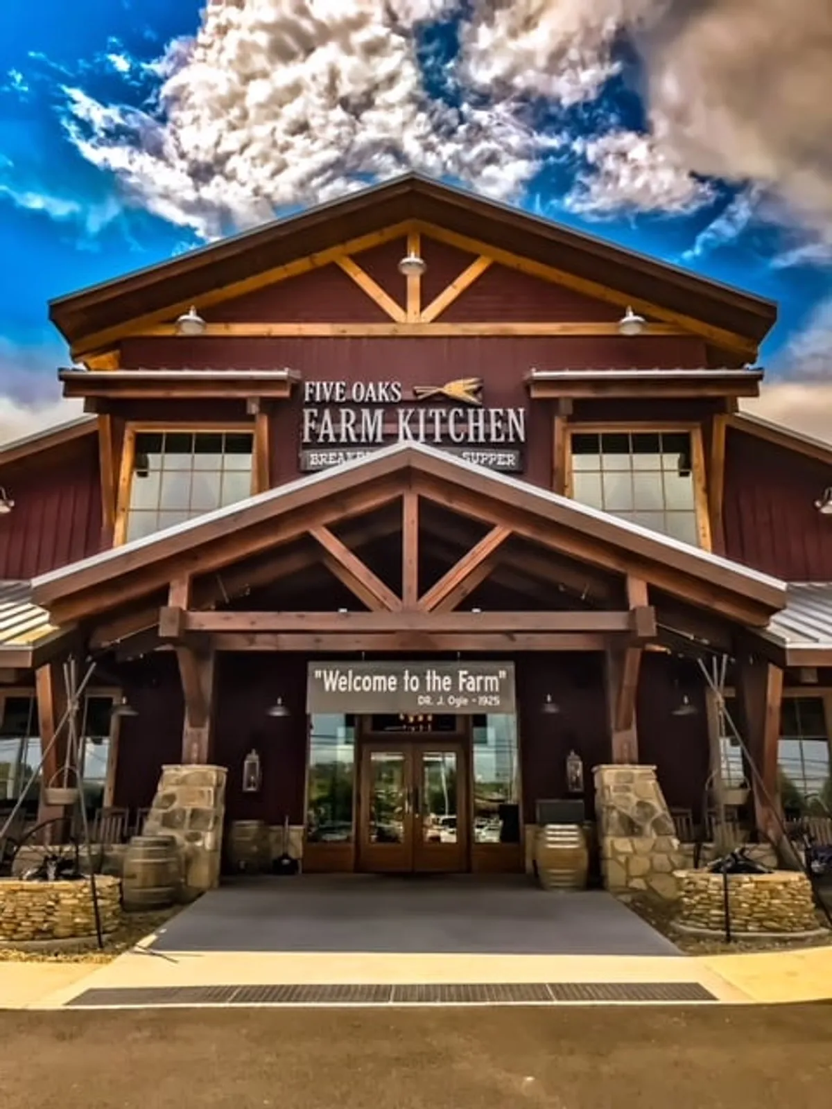 The exterior of a charming wooden structure marked 'Five Oaks Farm Kitchen,' featuring stone accents, outdoor seating, barrels, and clear signage. The space is lively and welcoming—ideal for dining or gathering after a day of adventure.