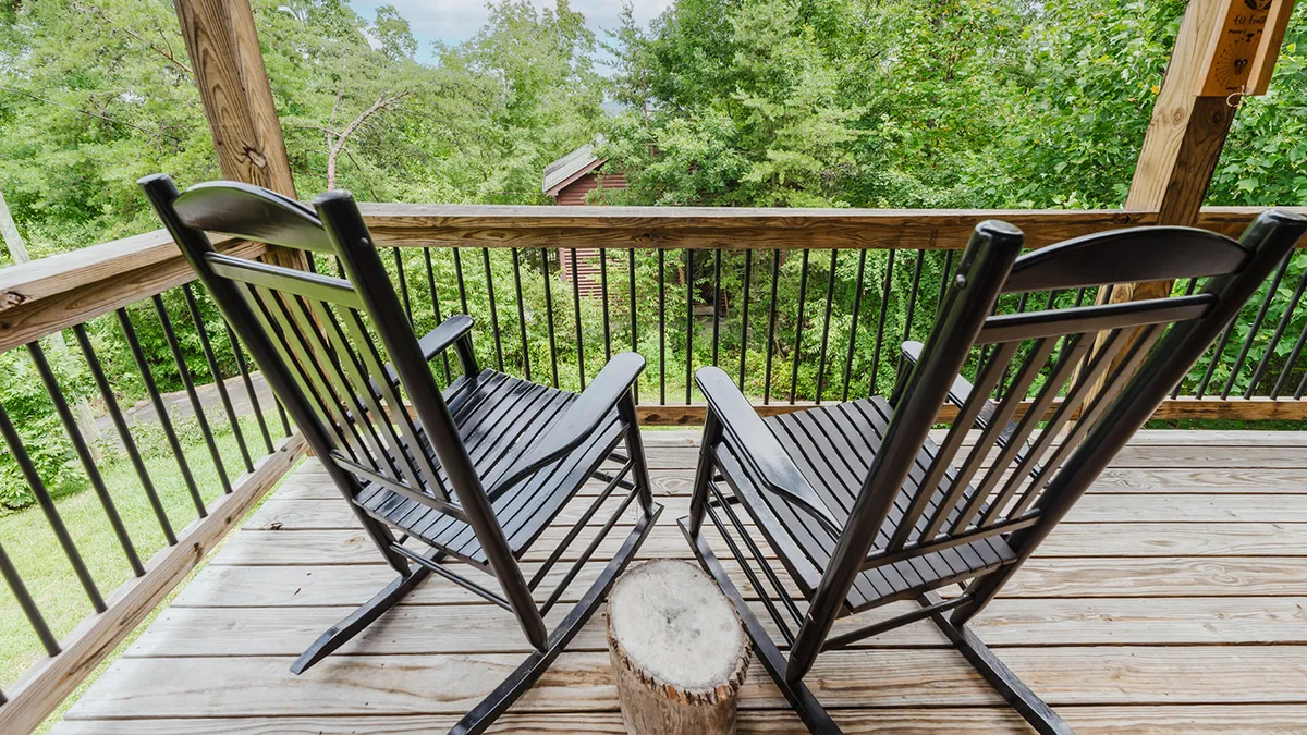 A cozy porch setting featuring rustic wooden rocking chairs, a stump table, and a wooden railing overlooking natural greenery. The soft lighting and inviting arrangement create a perfect spot for couples to relax together in the fresh mountain air.