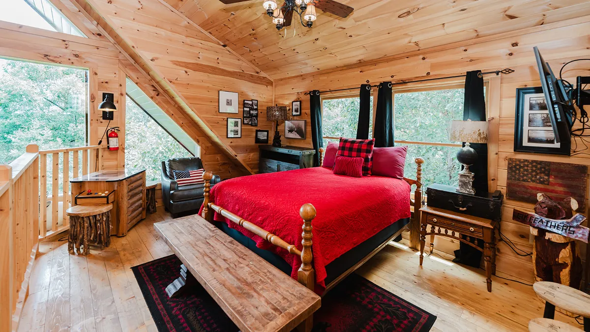 A cozy bedroom featuring a red bedspread, wooden furniture, large windows offering tranquil views of the forest, and decorative wall art. The room feels rustic and inviting, with soft natural light filtering through the windows and thoughtful decorative touches that add warmth.