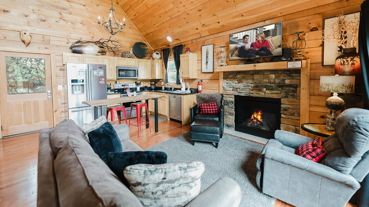 This image showcases a welcoming living room with a fireplace, plush sofa, kitchen area, and flat-screen TV. The space exudes warmth with its cozy seating, visible firelight, and large windows letting in natural light. The arrangement invites relaxation and togetherness, perfect for family gatherings or quiet evenings.
