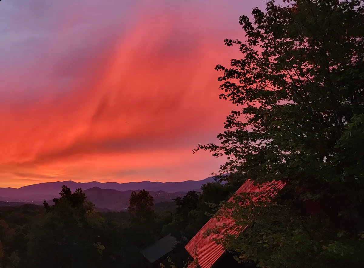 This image captures a vibrant sunset sky above distant mountains, with lush trees in the foreground and the rooftop of the house visible below. The tranquil setting invites guests to pause and enjoy nature’s beauty from a comfortable vantage point.