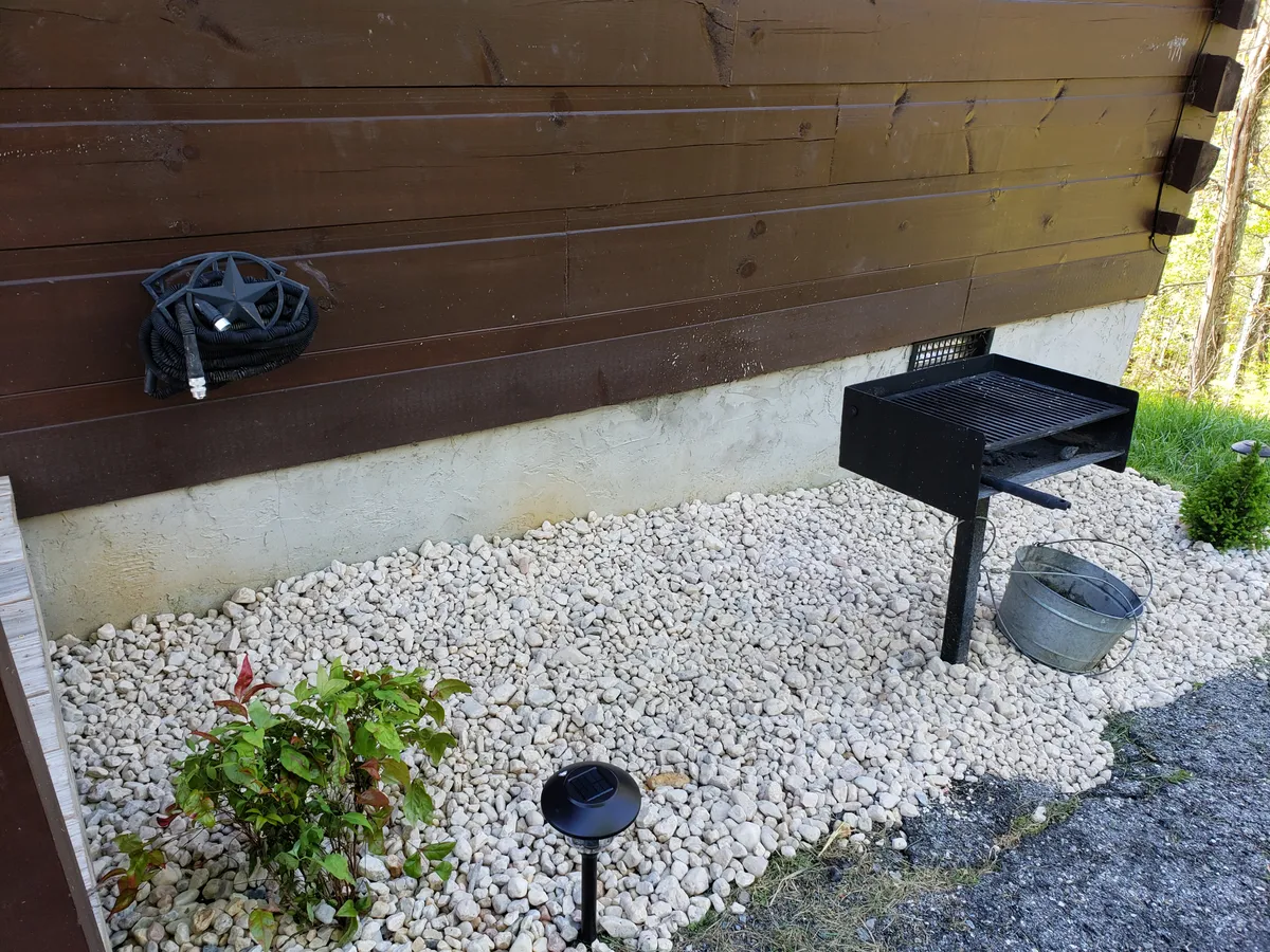 A close-up of an outdoor charcoal grill on a gravel area next to a cozy wooden railing and small potted plant. The scene invites thoughts of preparing a delicious barbecue in a relaxed, natural setting, perfect for gathering with friends or family after a day of exploration.