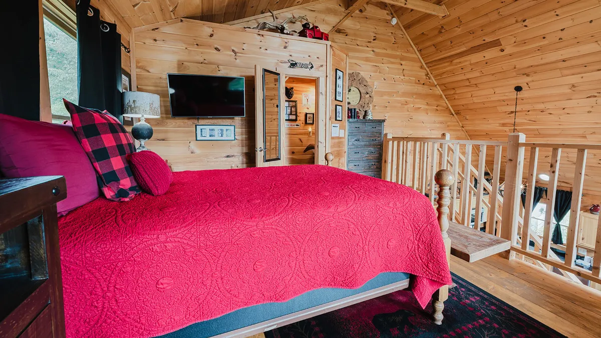 An inviting bedroom with a red quilted bedspread, wood-paneled walls, decorative shelving, a television, and a doorway leading to another room. The space radiates warmth, with thoughtful touches throughout and soft lighting creating a restful mood.