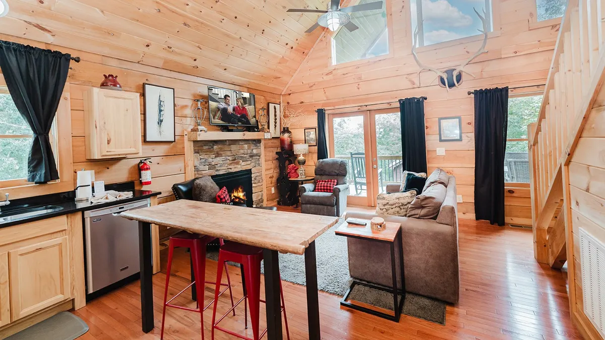 This image captures a warm, inviting interior with a fireplace, exposed wooden beams, a spacious kitchen island, large windows, and comfortable seating. The kitchen island is central, surrounded by stools, and the whole space is filled with natural light, creating an ambiance perfect for both cooking and communal dining.