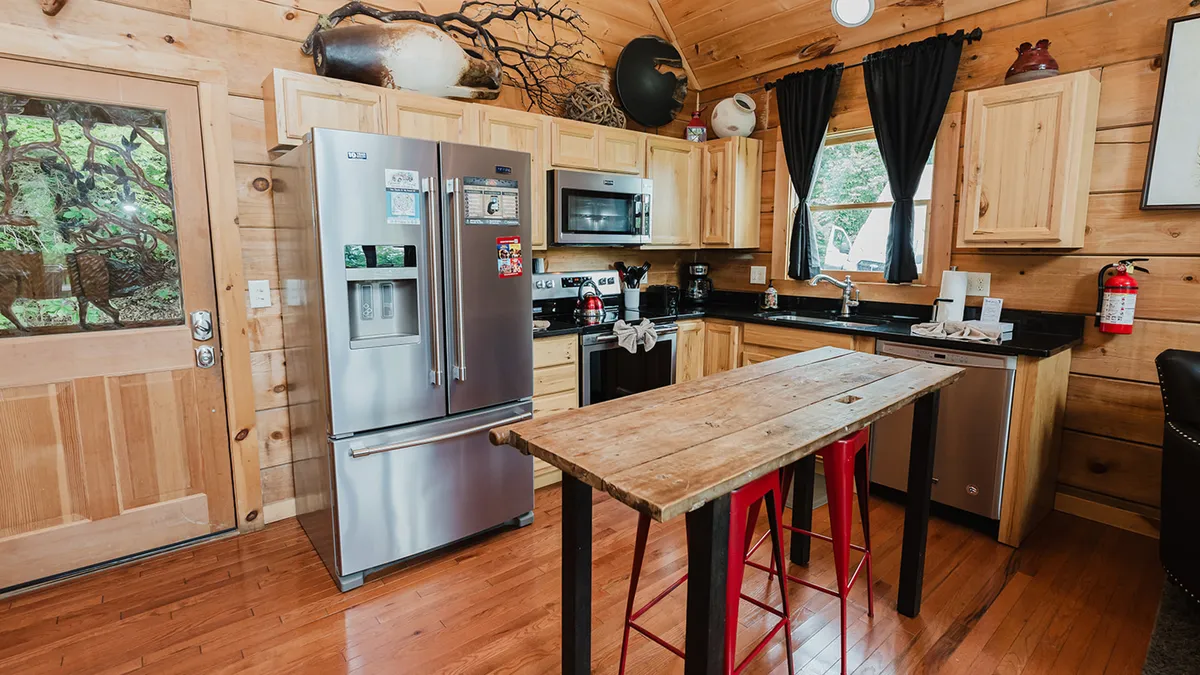 The scene spotlights a stainless steel refrigerator, wooden dining table, black countertops, and sunlit windows. There are decorative items on the shelves, creating a warm, rustic ambiance that still feels modern and practical for group cooking and dining activities.