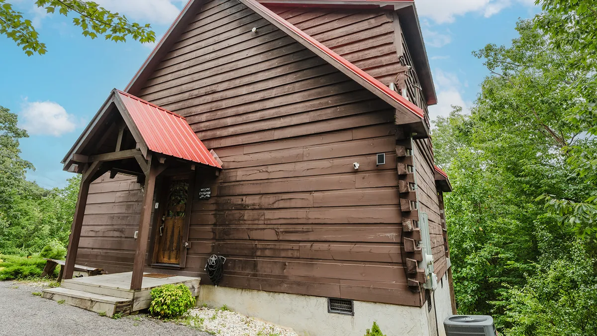 This image features the wooden exterior of the cabin with a rustic red roof, an inviting entrance porch, and lush surrounding greenery. The porch area is open and spacious, suggesting a welcoming spot to enjoy the outdoors, perhaps with a dog relaxing by your side.
