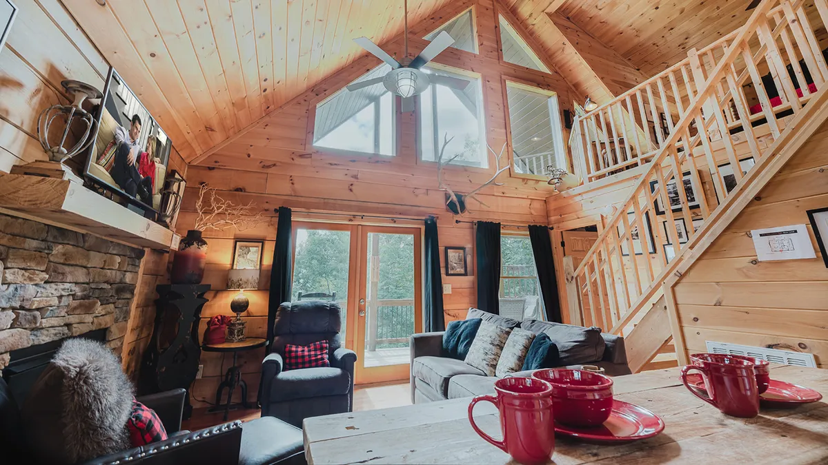 A spacious, bright cabin interior with a stone fireplace, large windows offering scenic views, a wooden staircase, and comfortable communal seating. The setup is perfect for relaxing, socializing, or enjoying after-adventure downtime with friends.